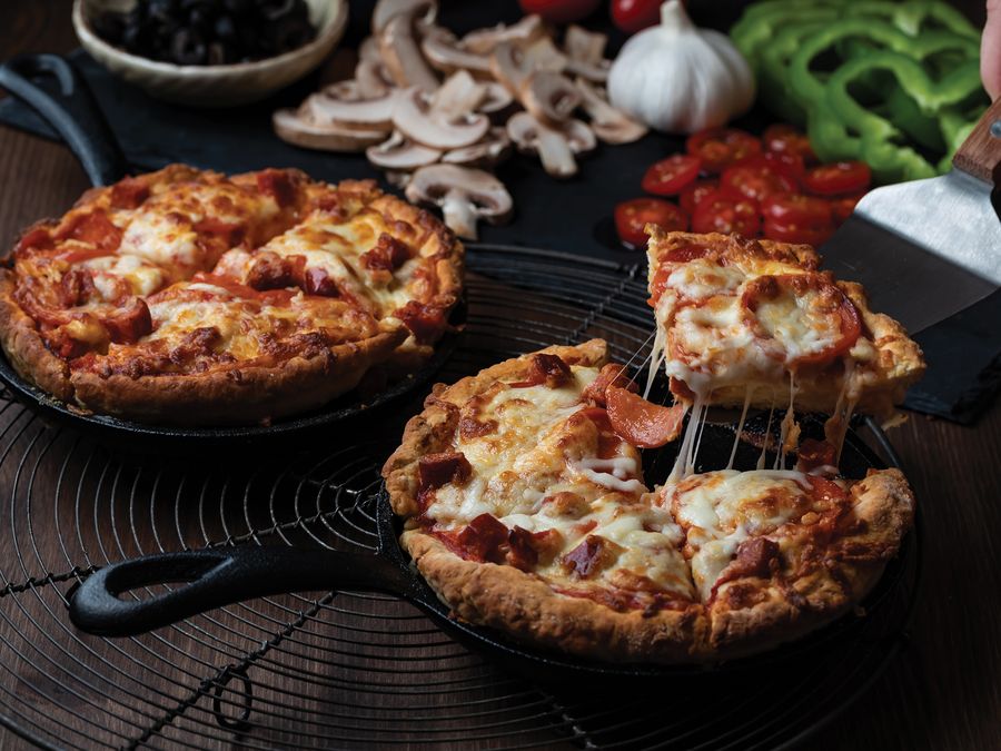 Two deep pan pepperoni pizzas in cast-iron pans, one slice lifted